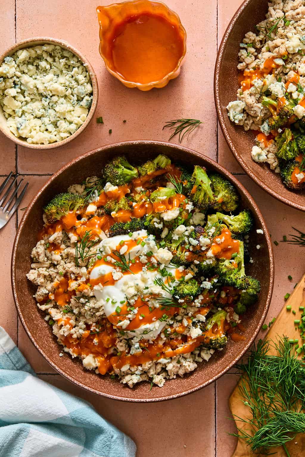 A bowl with scrambled turkey, roasted broccoli, a poached egg, blue cheese crumbles, and orange buffalo sauce drizzle. Blue cheese, sauce, and fresh herbs are on the side. A fork and napkin are nearby.