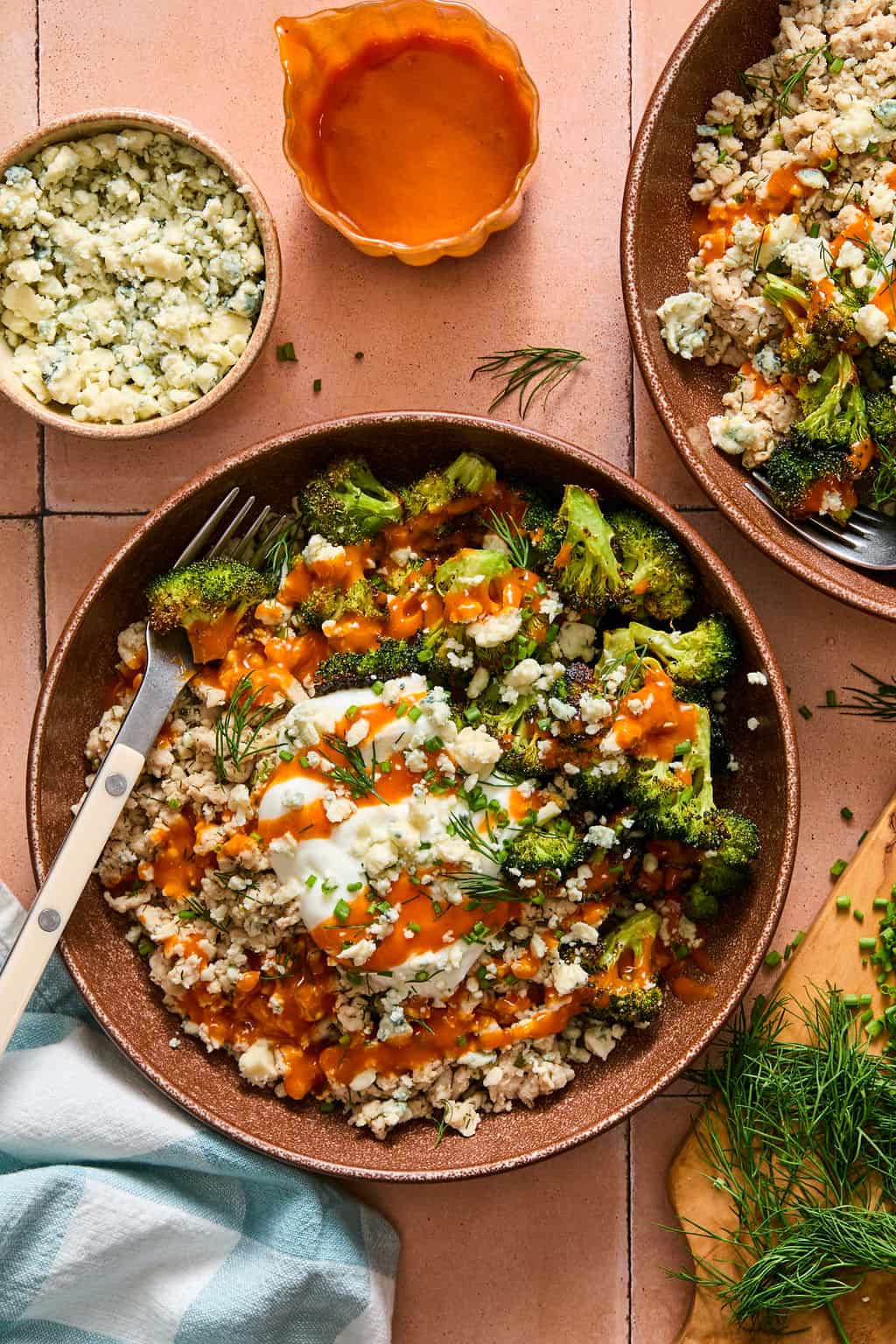 A bowl with cauliflower rice, roasted broccoli, a dollop of yogurt, crumbled blue cheese, fresh herbs, and orange sauce. A fork rests in the bowl. Nearby are a plate, a small bowl of cheese, and a dish of sauce.