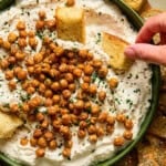 A bowl of creamy dip topped with roasted chickpeas and herbs. A hand dips a piece of toasted bread into the bowl, with more pieces of bread surrounding the bowl on the side.
