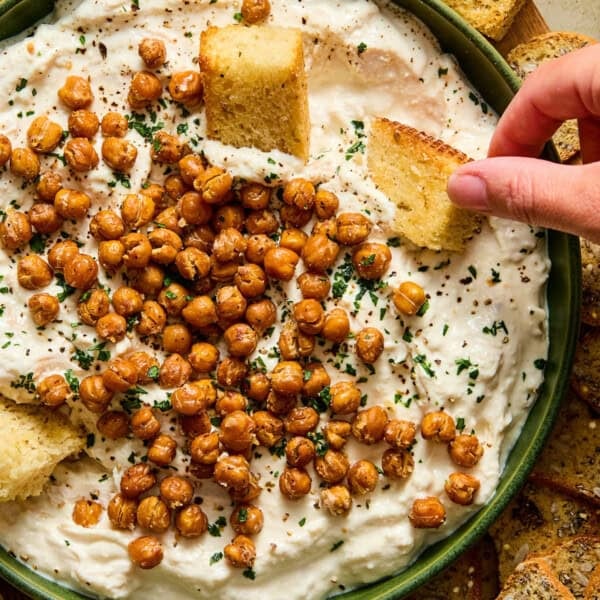 A bowl of creamy dip topped with roasted chickpeas and herbs. A hand dips a piece of toasted bread into the bowl, with more pieces of bread surrounding the bowl on the side.