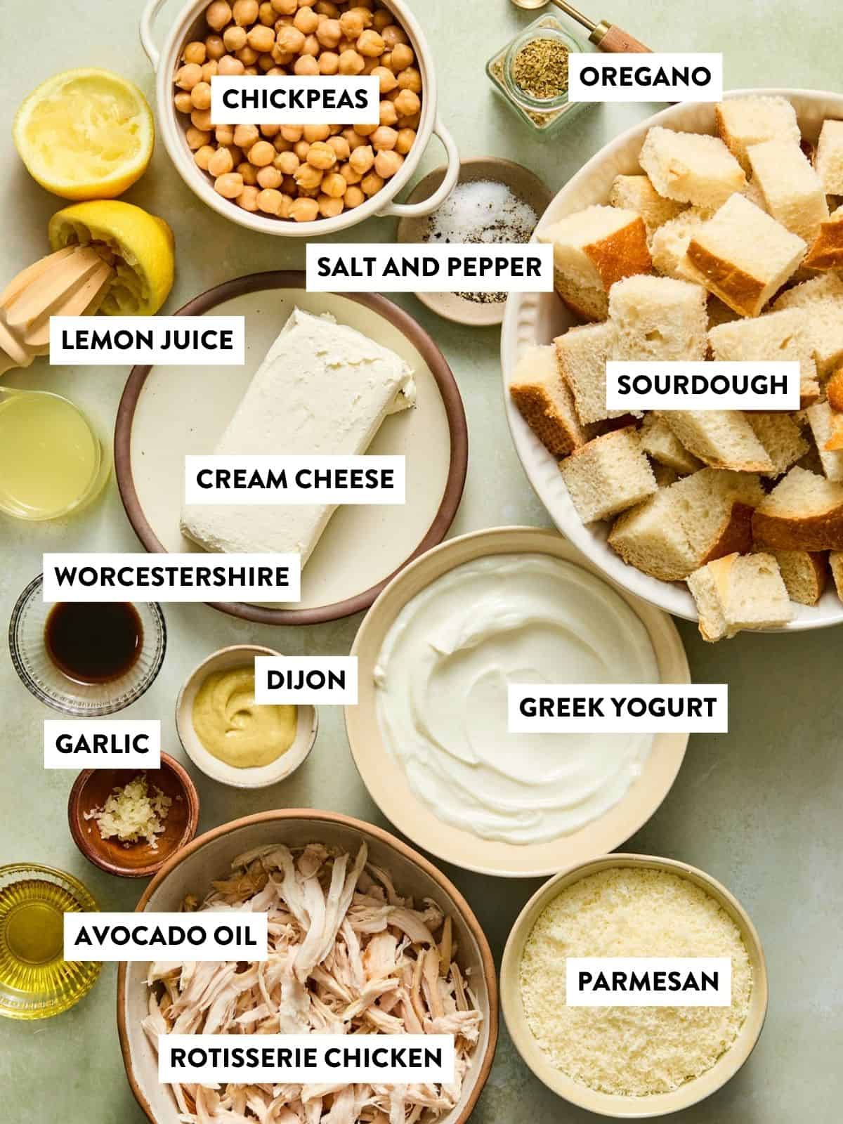 Various ingredients in bowls and plates labeled: chickpeas, oregano, sourdough bread, lemon juice, salt and pepper, cream cheese, Worcestershire, Dijon, Greek yogurt, garlic, avocado oil, rotisserie chicken, and parmesan.