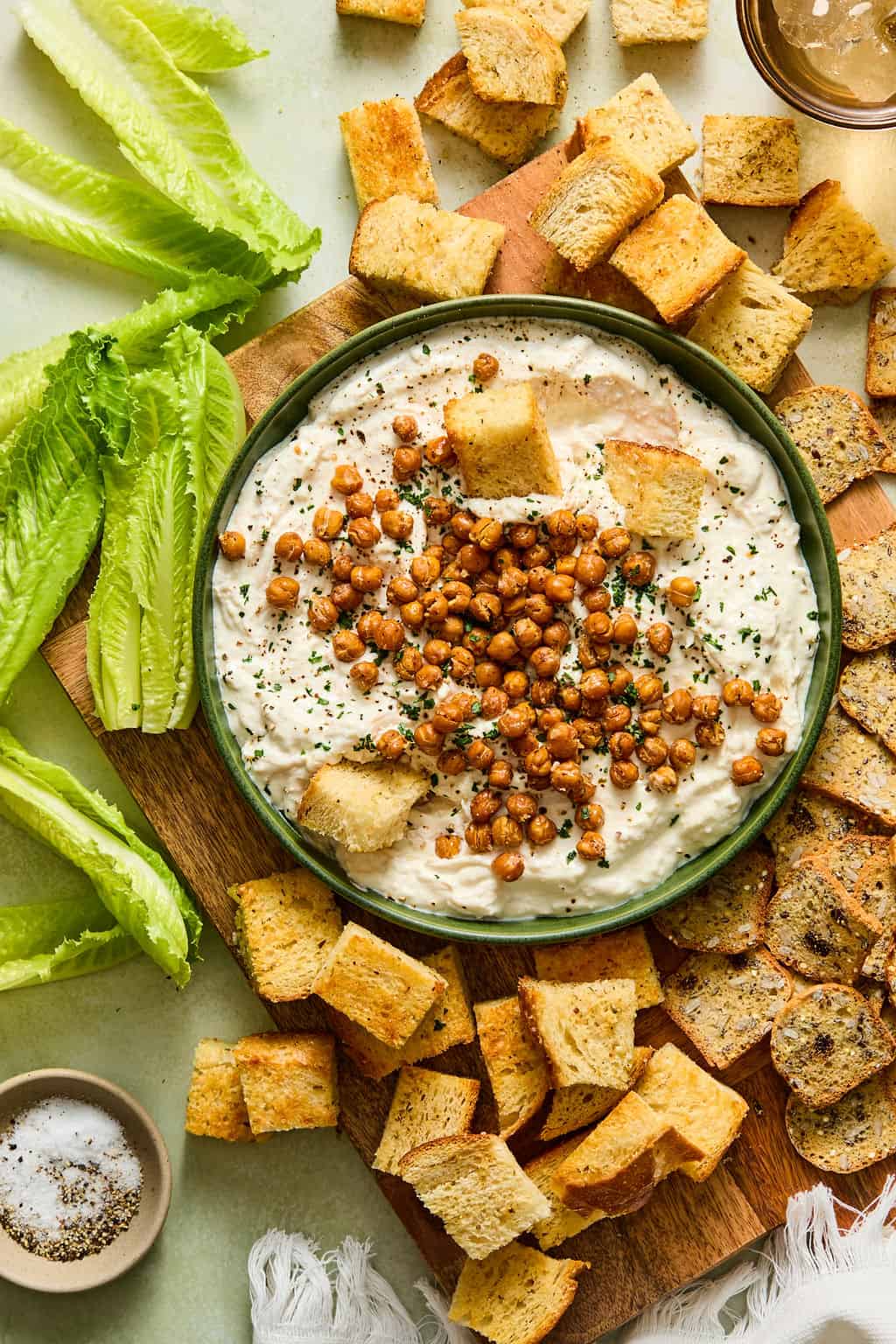 A bowl of creamy chicken caesar dip topped with roasted chickpeas, surrounded by lettuce leaves, croutons, and toasted bread slices on a wooden board. A small bowl of salt is nearby.