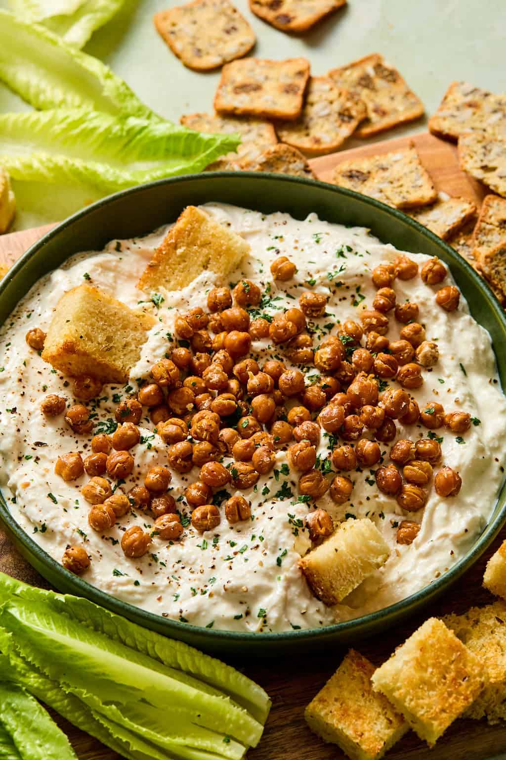 A bowl of creamy dip topped with roasted chickpeas, chopped herbs, and croutons, surrounded by lettuce leaves and assorted crackers.