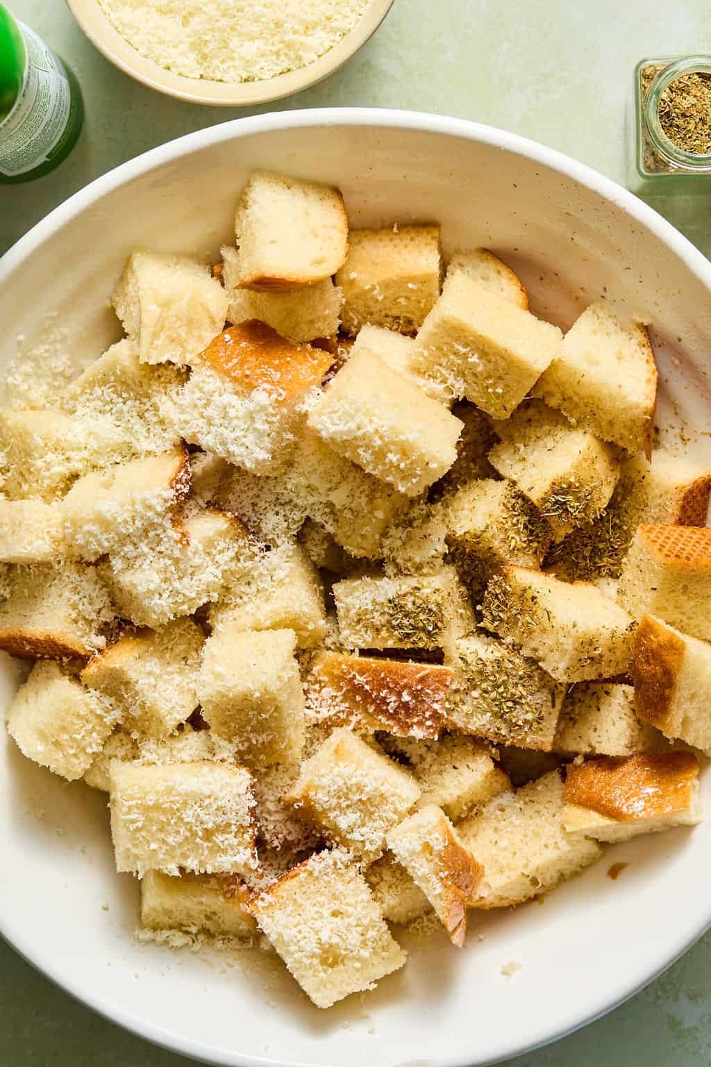 Cubed bread pieces in a white bowl, sprinkled with grated cheese and dried herbs, ready to be tossed. A bowl of cheese and a spice container are nearby.
