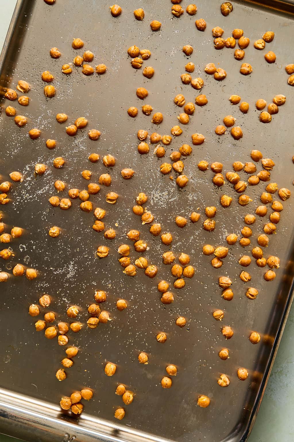 Scattered roasted chickpeas on a dark metal baking sheet, sprinkled with salt.