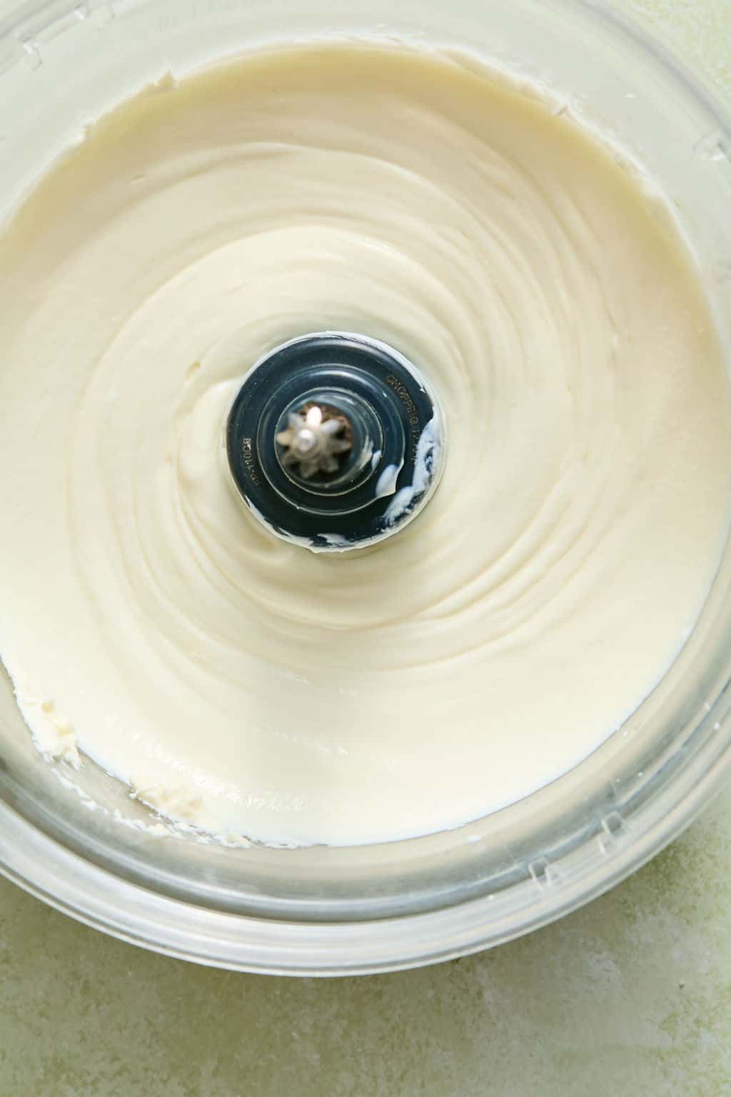 A food processor bowl filled with smooth, creamy greek yogurt and cream cheese viewed from above. The metal blade is visible at the center of the blended contents.