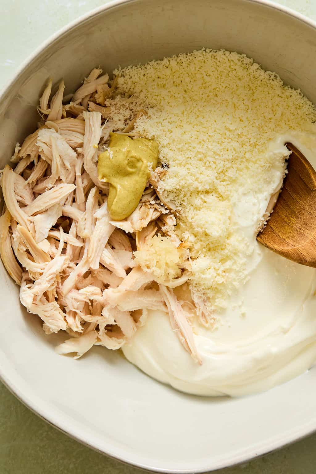 A bowl with shredded chicken, grated cheese, yogurt, mustard, and a wooden spoon, ready to be mixed together.