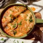 A bowl of creamy sausage tortellini soup with herbs, and shredded cheese on top, served on a wooden board in bright sunlight.