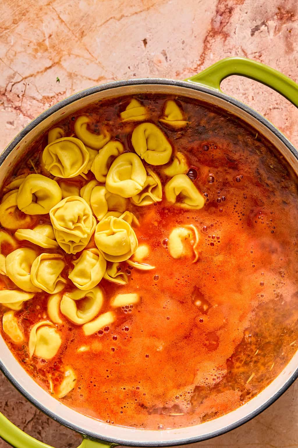 A pot of soup with cheese tortellini being added, viewed from above on a marble countertop.