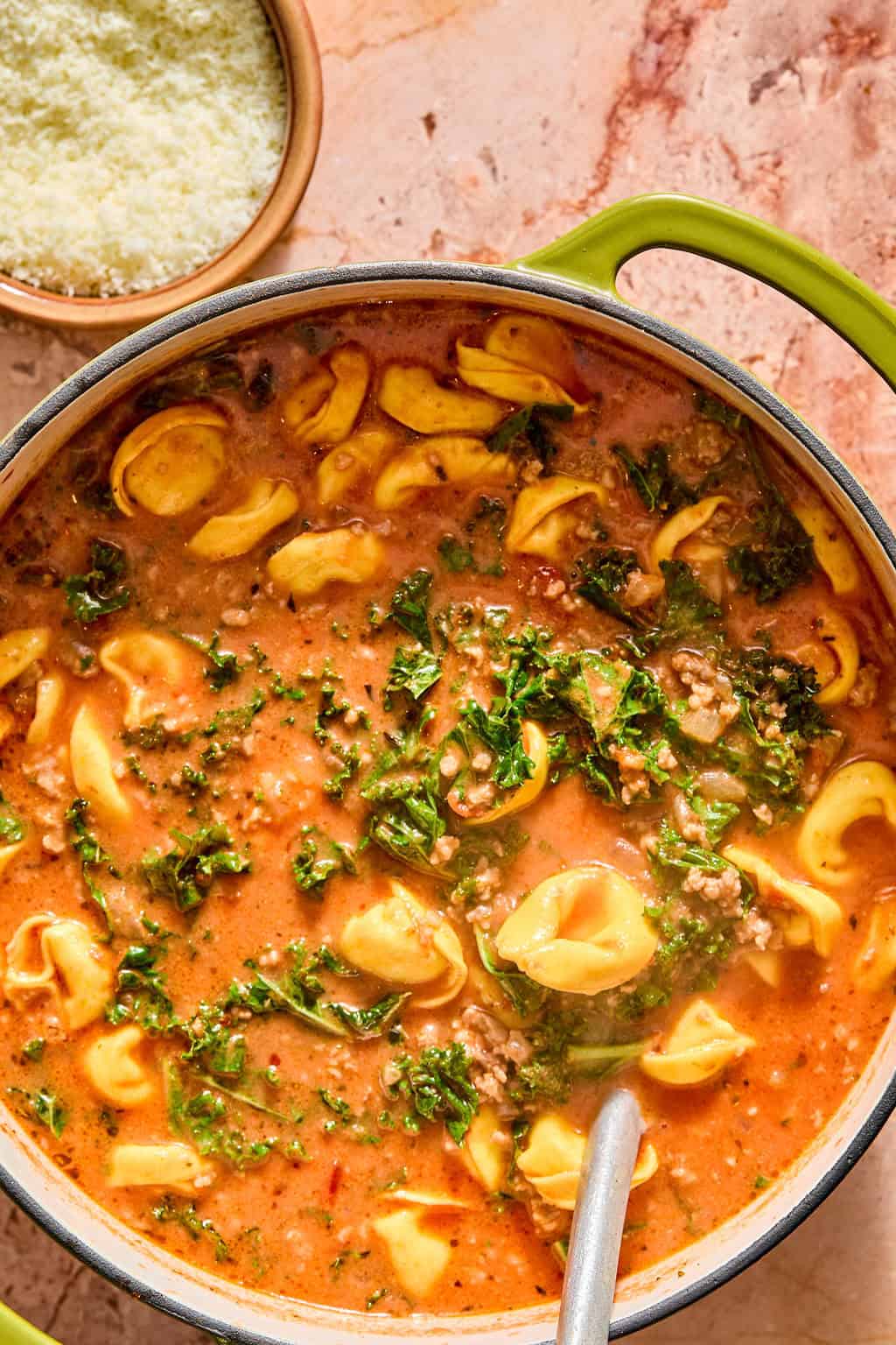 A pot of creamy tomato tortellini soup with ground meat and kale, next to a bowl of grated cheese. A spoon rests in the pot.