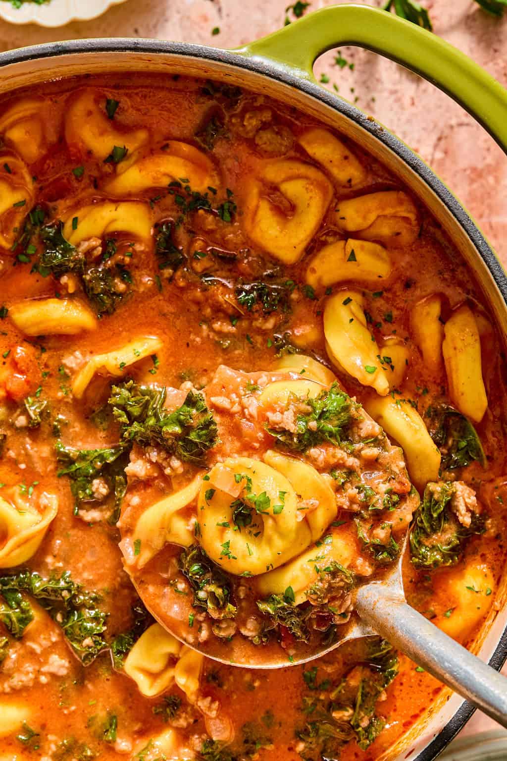 A pot of creamy sausage and tortellini soup with ground meat, kale, and a tomato-based broth. A ladle lifts a serving, showing stuffed pasta, chopped greens, and herbs in the rich, hearty soup.