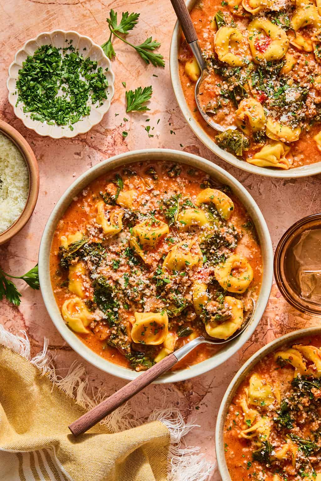 Three bowls of creamy tomato tortellini soup with kale, topped with grated cheese and chopped herbs. A bowl of grated cheese, herbs, and a drink are nearby on a textured surface with a beige cloth.