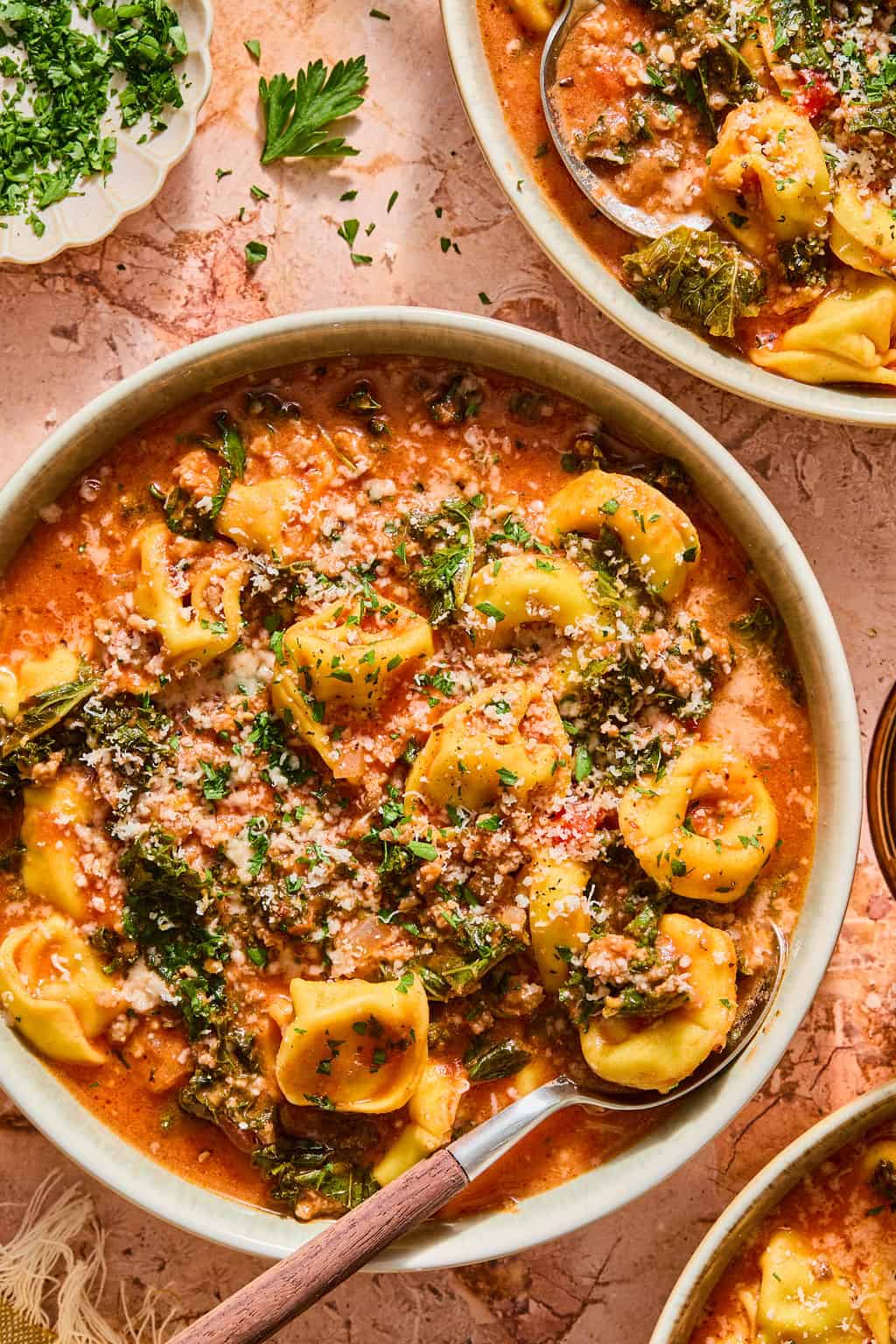 Bowl of creamy tortellini soup with sausage, kale, and herbs, topped with grated cheese and parsley. A spoon rests in the bowl, and fresh herbs are visible on the side.