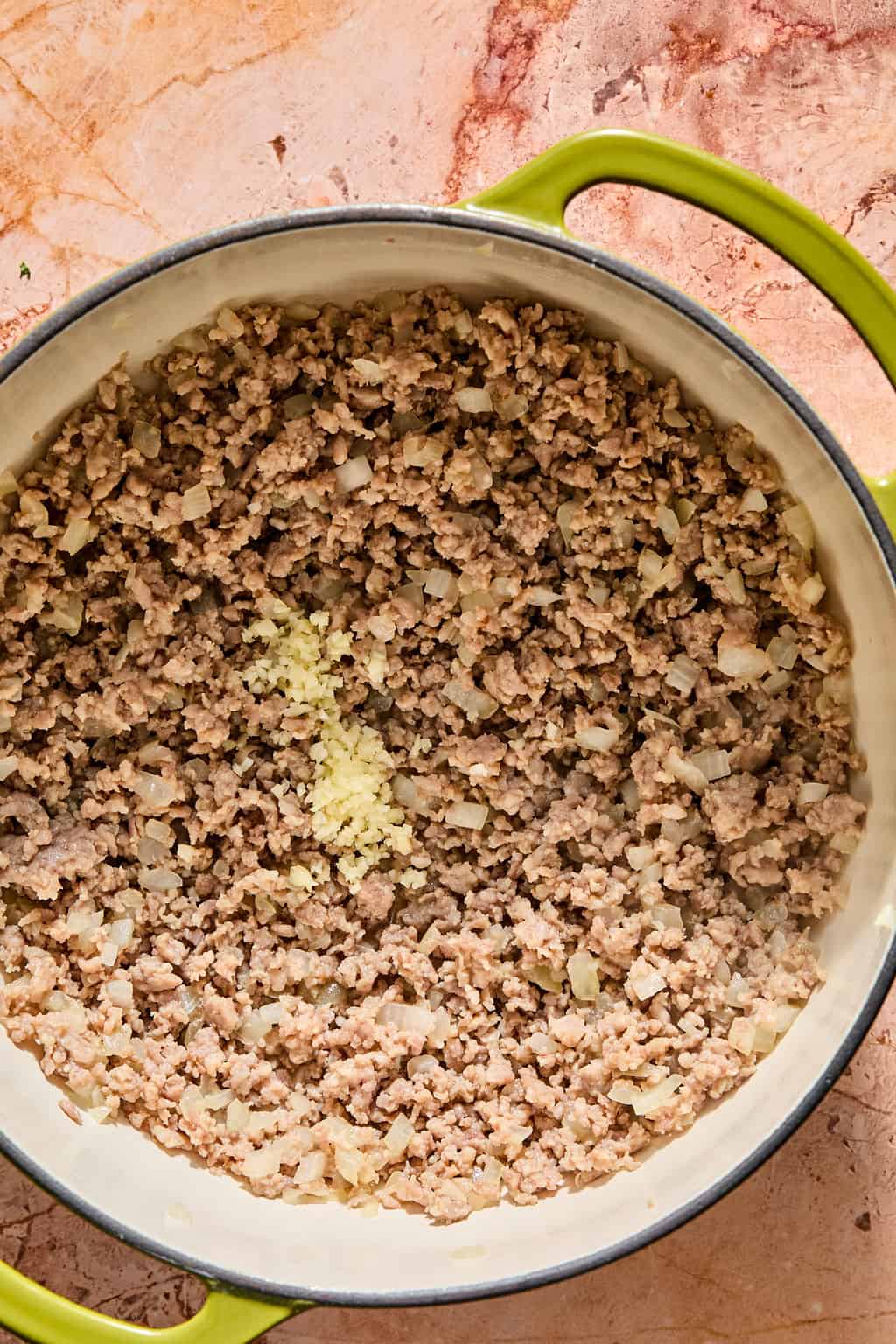 Ground meat and diced onions cooking in a large green pot, with a small pile of minced garlic added to the center.