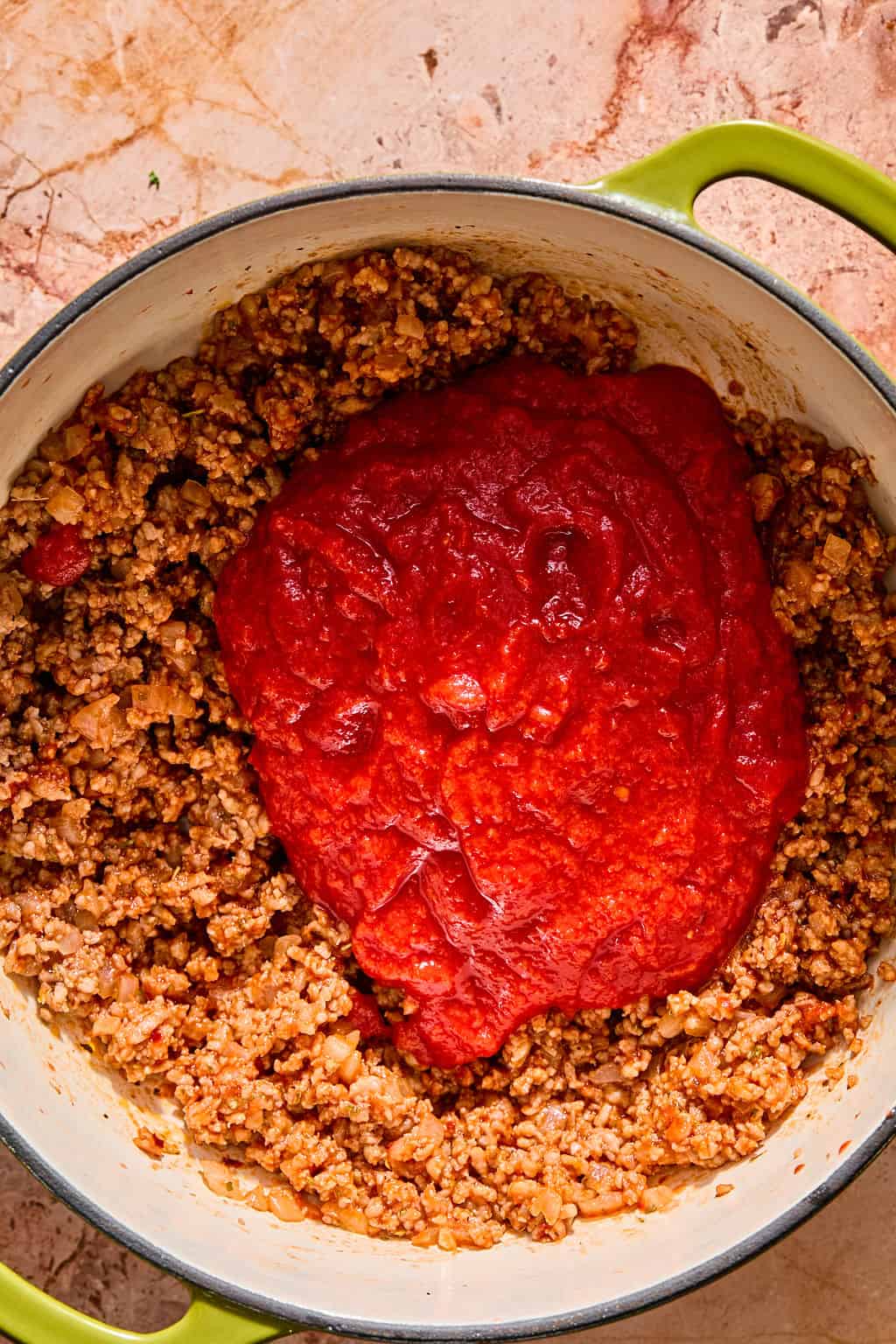 Ground meat and vegetables cooked in a pot with a large portion of tomato sauce on top, ready to be mixed, on a light brown countertop.