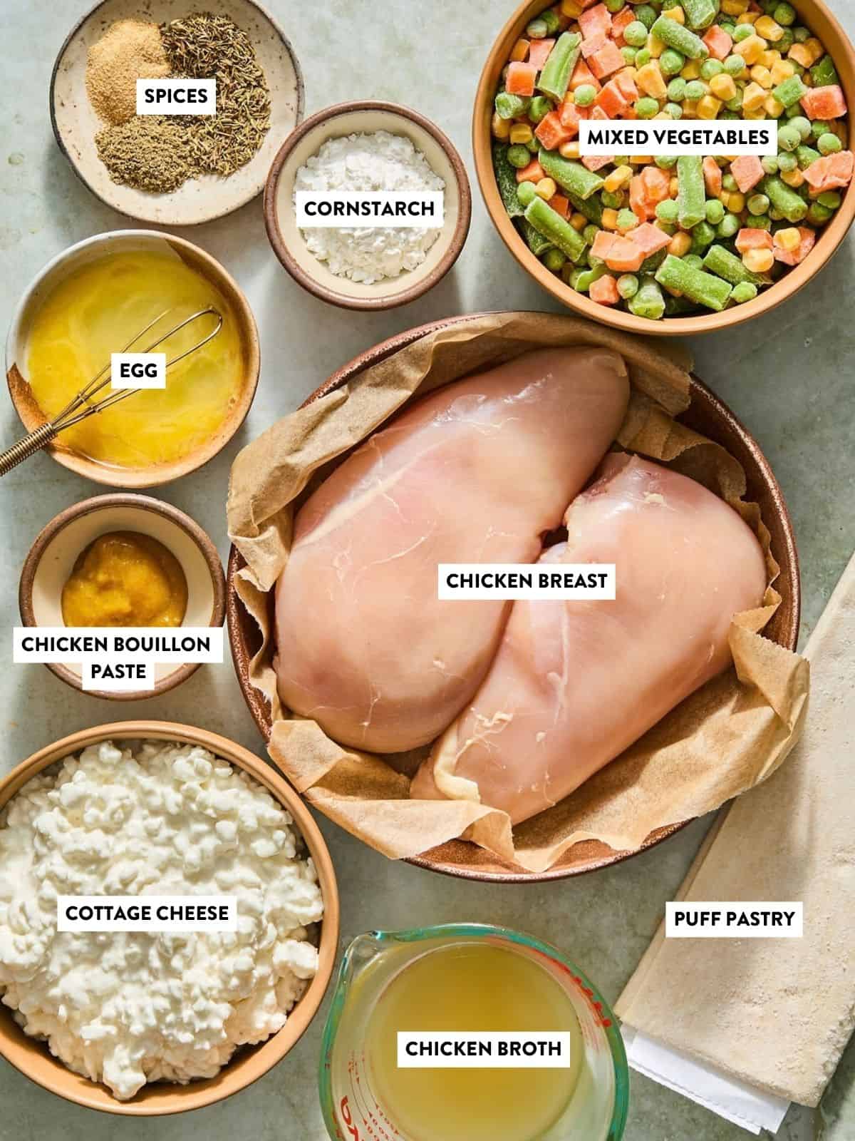 Top-down view of labeled ingredients for a recipe: spices, cornstarch, mixed vegetables, egg, chicken bouillon paste, chicken breast, cottage cheese, puff pastry, and chicken broth, all arranged on a table.
