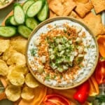 A bowl of creamy fried pickle dip topped with toasted breadcrumbs, pickles, and dill surrounded by sliced cucumbers, potato chips, crackers, and sliced mini peppers on a wooden platter.
