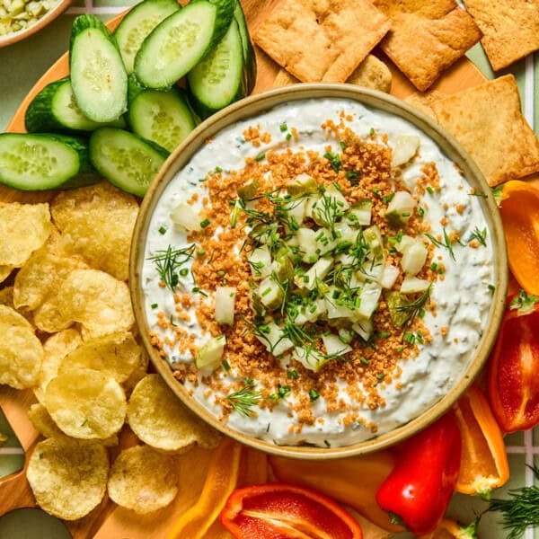 A bowl of creamy fried pickle dip topped with toasted breadcrumbs, pickles, and dill surrounded by sliced cucumbers, potato chips, crackers, and sliced mini peppers on a wooden platter.