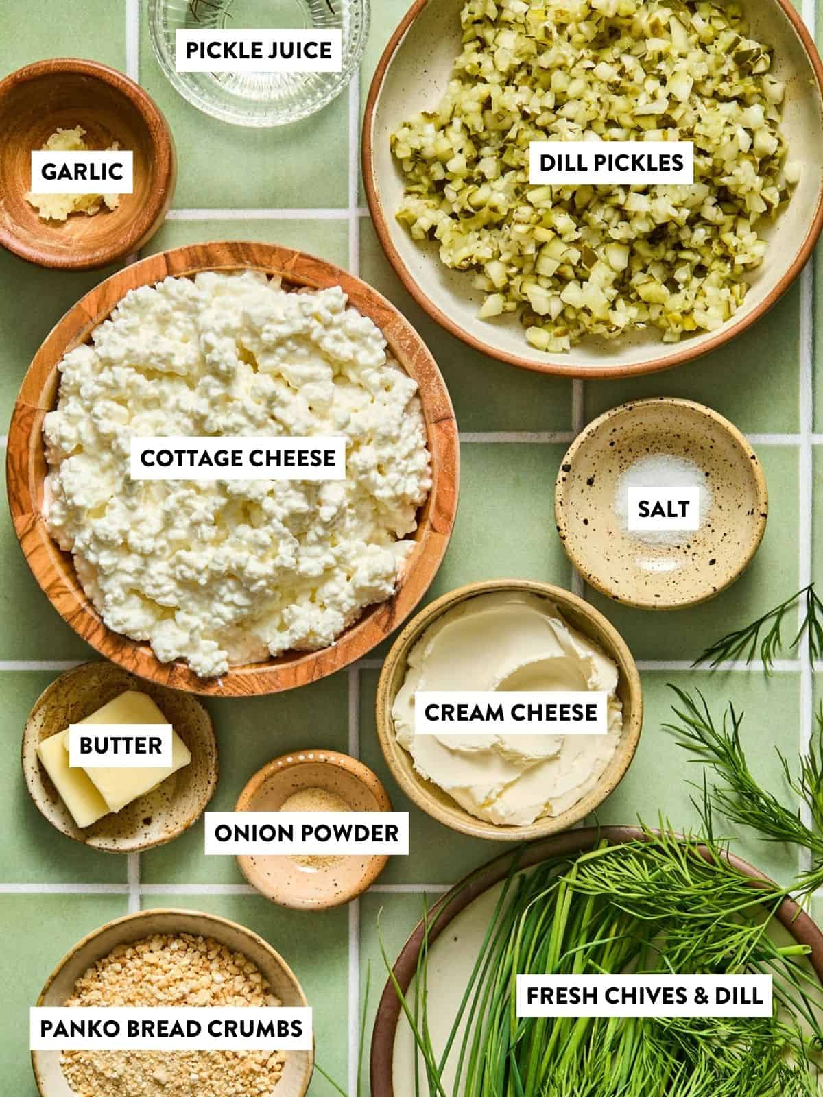 Ingredients arranged in bowls on a green tile surface, including cottage cheese, cream cheese, chopped dill pickles, garlic, butter, panko bread crumbs, salt, onion powder, pickle juice, and fresh chives with dill.