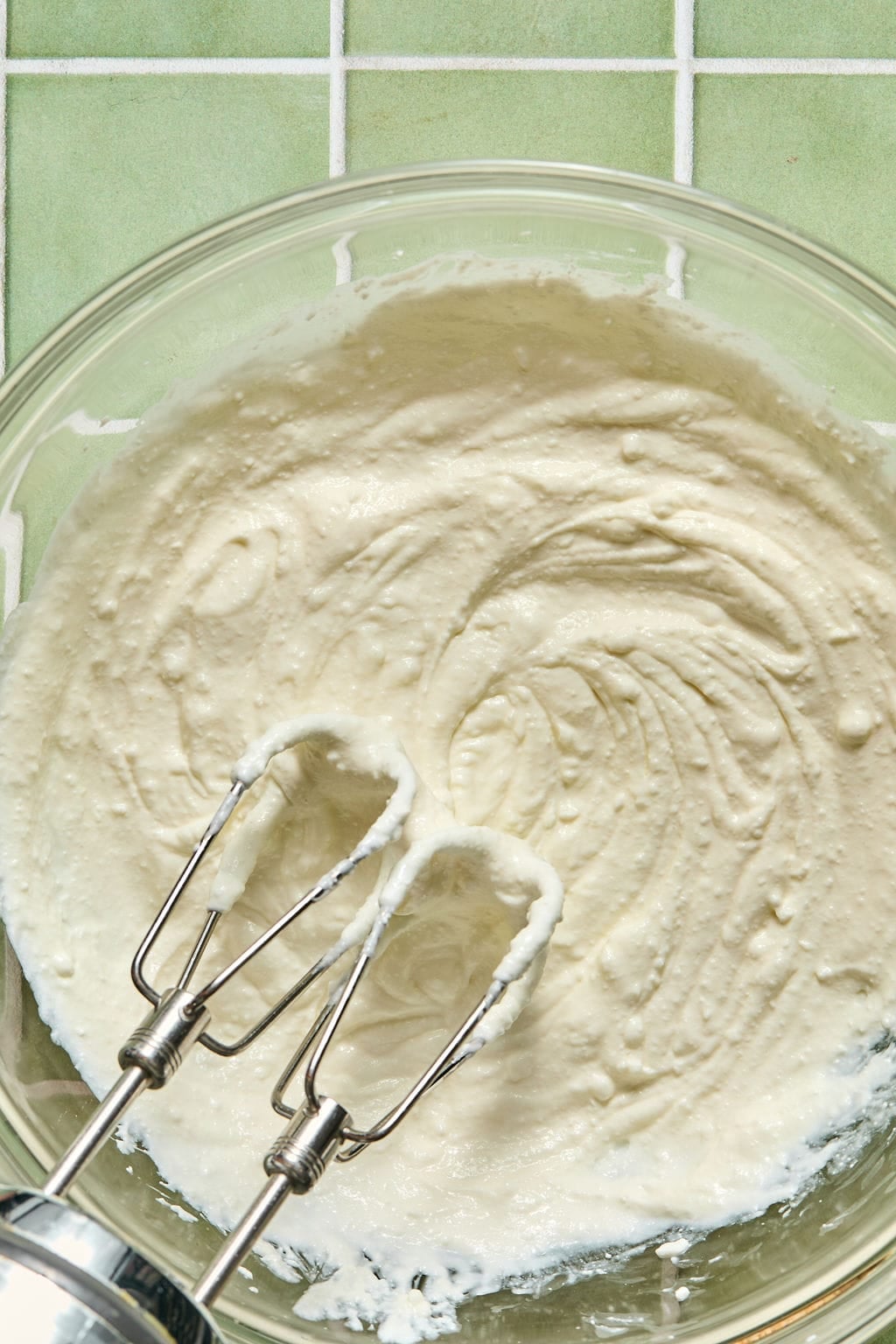 A glass bowl filled with creamy dip sits on a green tiled surface, with two metal beaters resting inside, coated in the mixture.