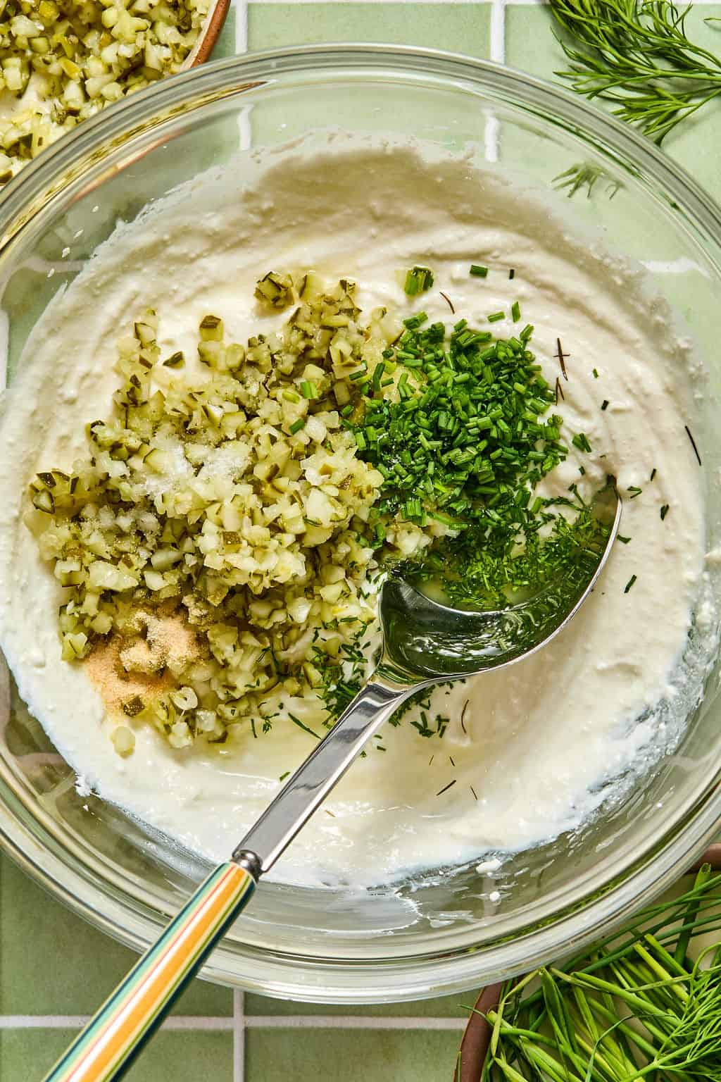 A bowl of creamy white dip with chopped pickles, chives, dill, and spices on top. A spoon rests in the bowl, ready to mix the ingredients. Fresh dill and a small bowl of pickles are in the background.