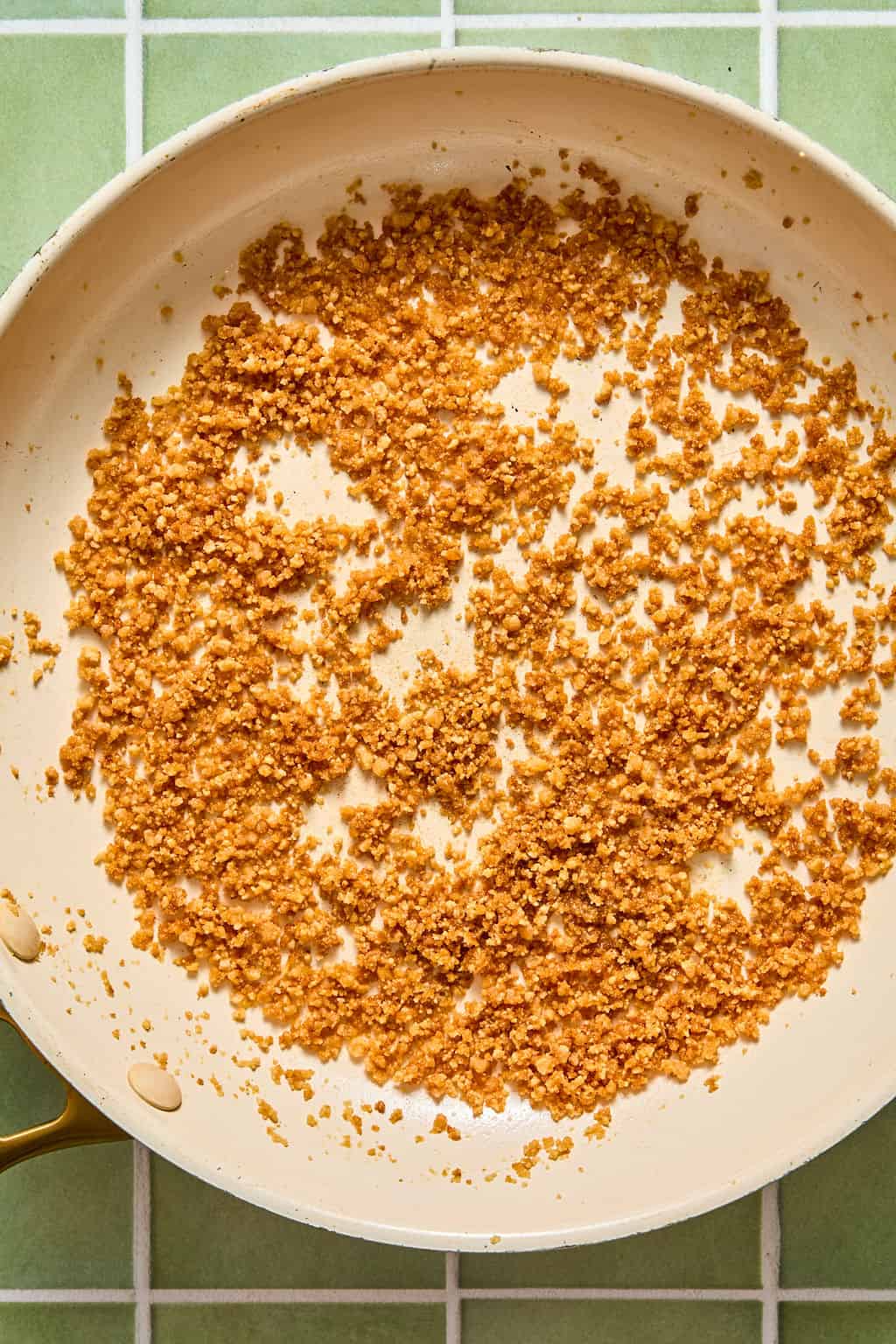 Golden brown toasted breadcrumbs spread in an even layer in a light-colored frying pan on a green and white tiled surface.
