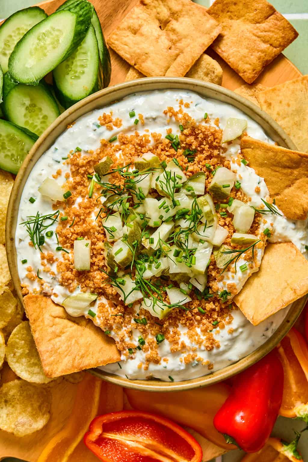 A bowl of fried dill pickle dip topped with chopped pickles, herbs, and crunchy breadcrumbs, surrounded by pita chips, cucumber slices, potato chips, and red bell pepper slices.