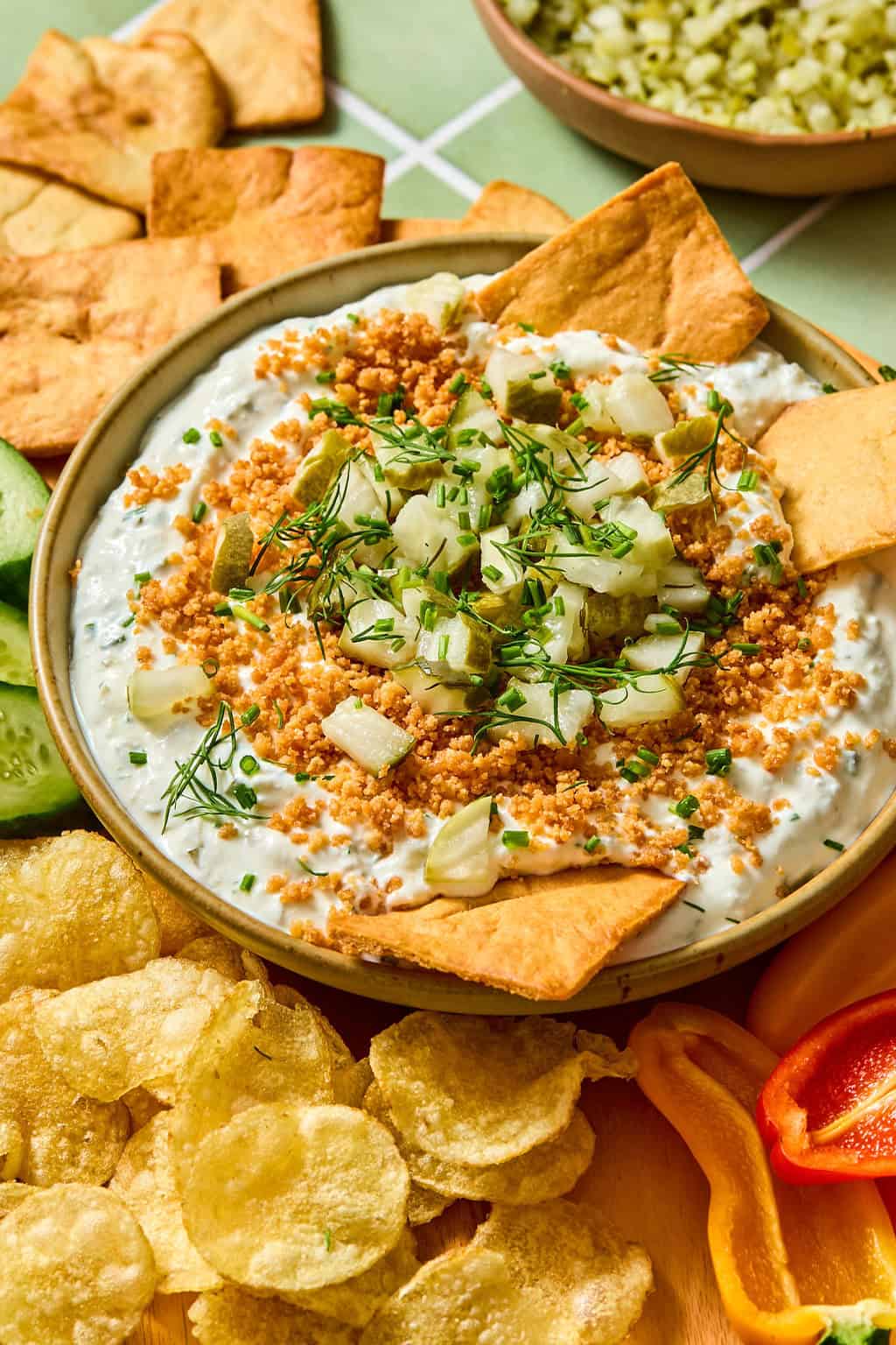 A bowl of creamy dip topped with chopped pickles, herbs, and crumbs, surrounded by pita chips, potato chips, cucumber slices, and bell pepper slices.