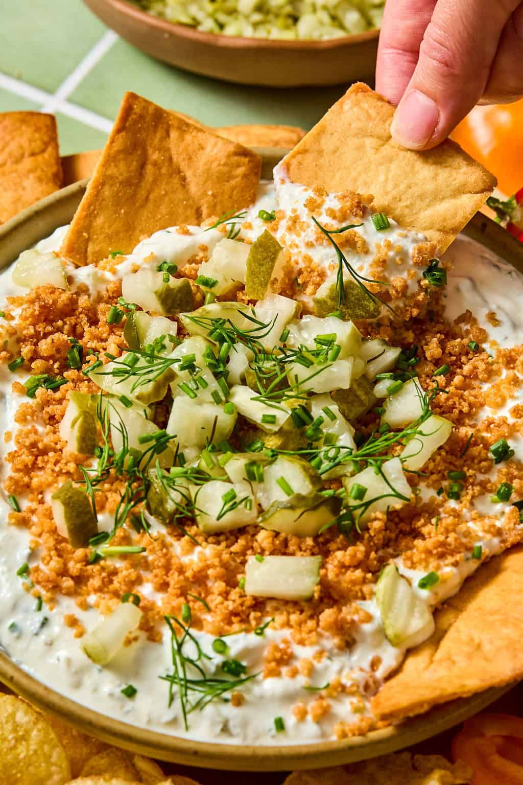 A hand dips a pita chip into a creamy dip topped with chopped pickles, fresh herbs, and crunchy crumbs, surrounded by more pita chips and some chips on the side.
