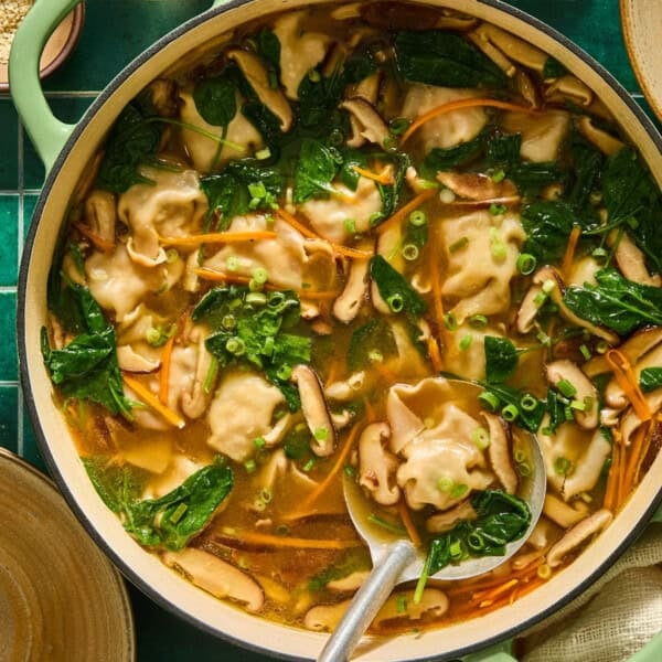 A pot of homemade dumpling soup with spinach, sliced mushrooms, carrots, chopped green onions, and a clear broth. A large spoon rests in the pot, ready to serve.