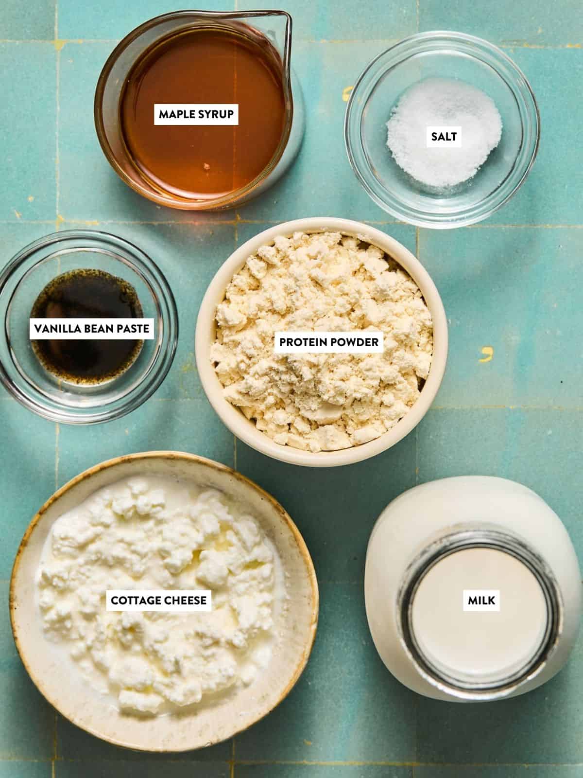 Six labeled ingredients on a teal surface—maple syrup, salt, vanilla bean paste, protein powder, cottage cheese, and milk—each in separate bowls or containers, ready to be blended into a creamy Ninja Creami protein ice cream.