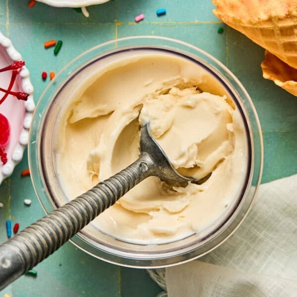 A metal ice cream scoop in a bowl of vanilla ninja creami protein ice cream sits on a green surface, with part of a waffle cone, sprinkles, cherries, and a white napkin nearby.