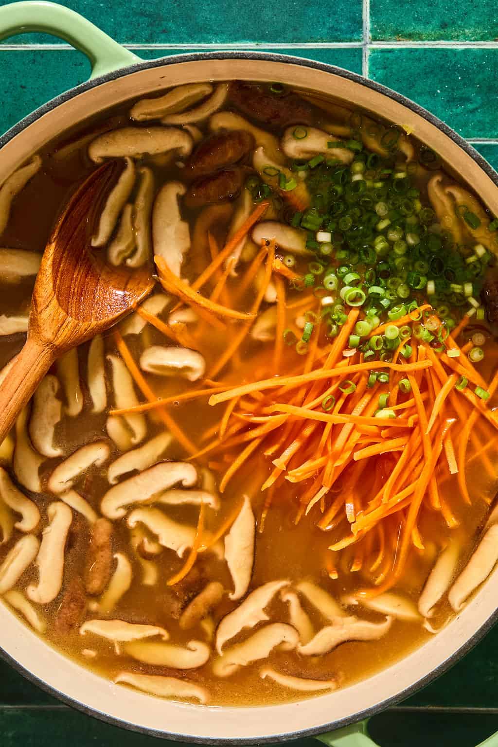 A pot of soup with sliced mushrooms, julienned carrots, chopped green onions, and a wooden spoon, on a green tiled surface.