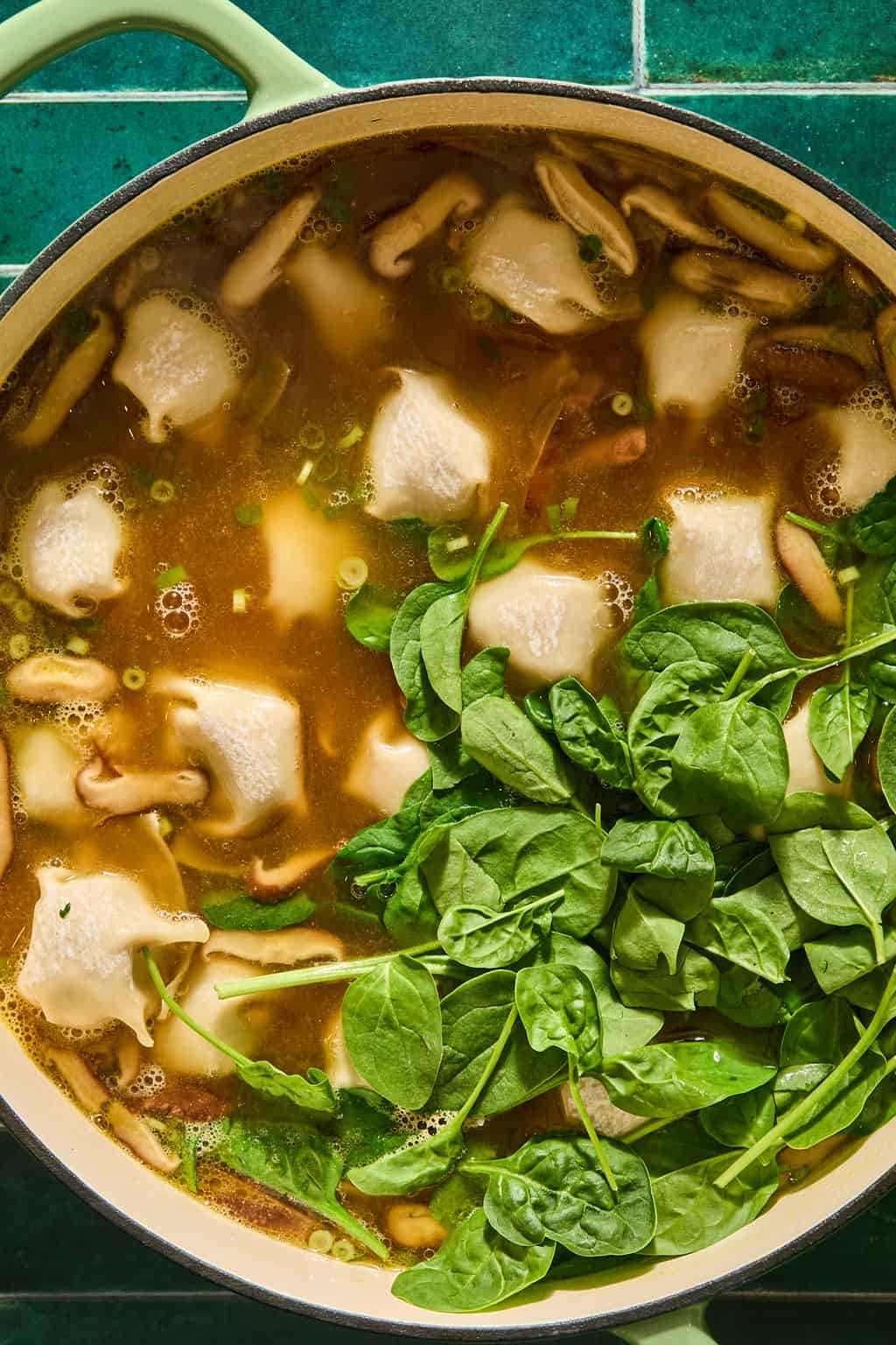 A pot of soup with dumplings, sliced mushrooms, and fresh spinach leaves cooking in broth, shown from above on a green tiled surface.