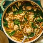 A pot of soup with dumplings, spinach, mushrooms, and carrots sits on a green tile surface. A ladle holds a dumpling, and bowls of chopped green onions and other toppings are nearby.