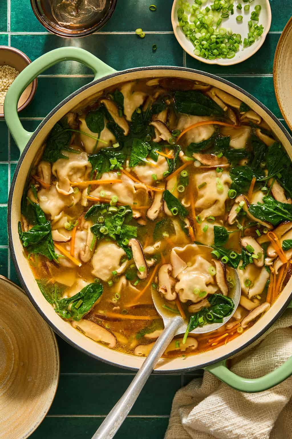 A pot of soup with dumplings, spinach, mushrooms, and carrots sits on a green tile surface. A ladle holds a dumpling, and bowls of chopped green onions and other toppings are nearby.