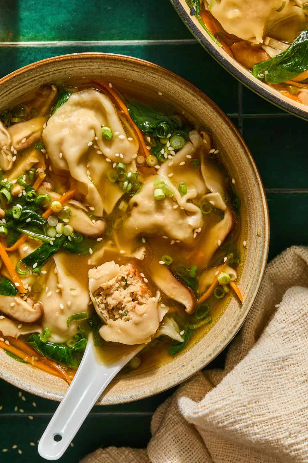 A bowl of dumpling soup with sliced mushrooms, spinach, carrots, and green onions, topped with sesame seeds. A soup spoon holds a dumpling, revealing a meat and vegetable filling.