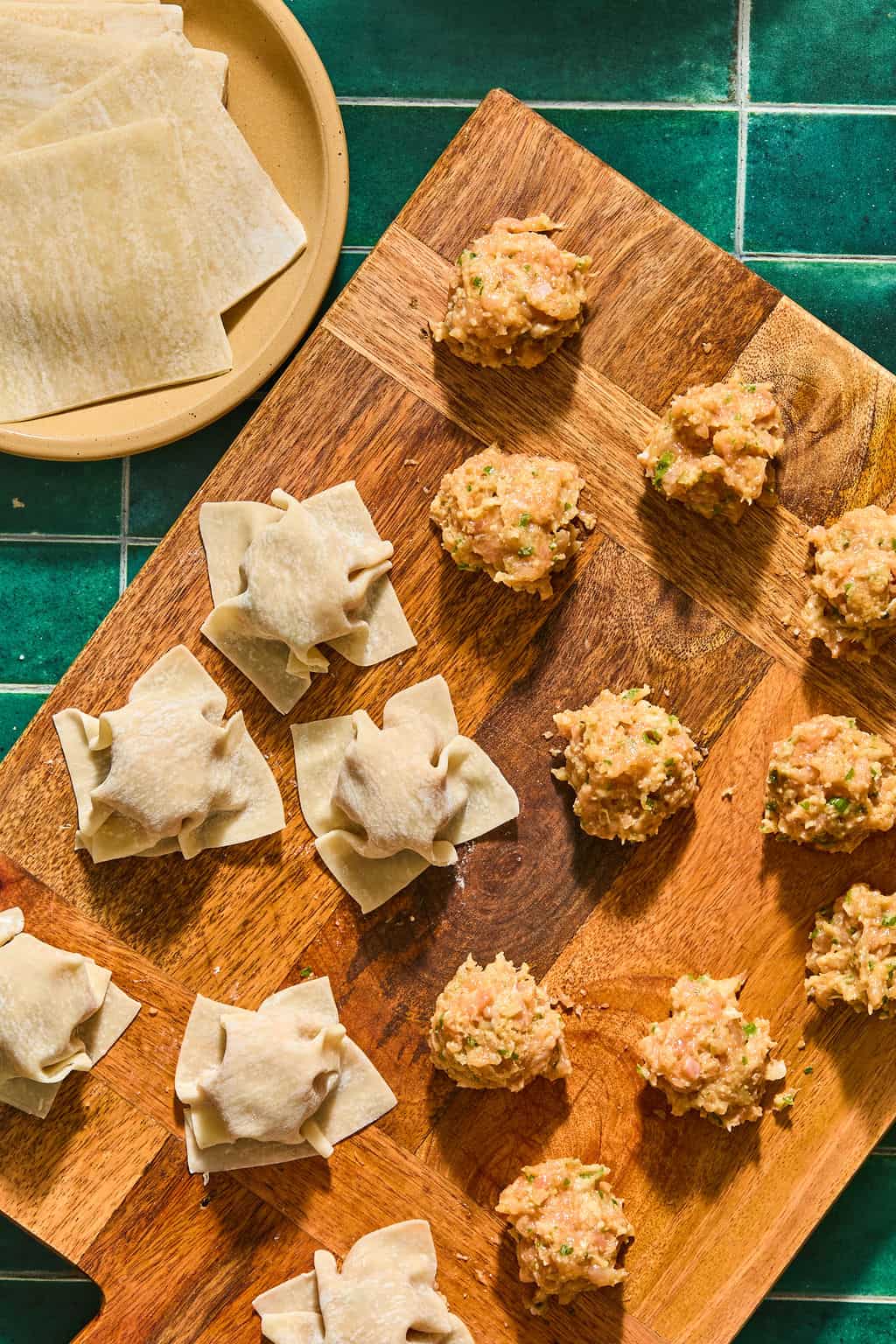 A wooden board holds scoops of a meat filling and partially folded wontons. Square wonton wrappers are stacked on a nearby plate, all on a green tiled surface.