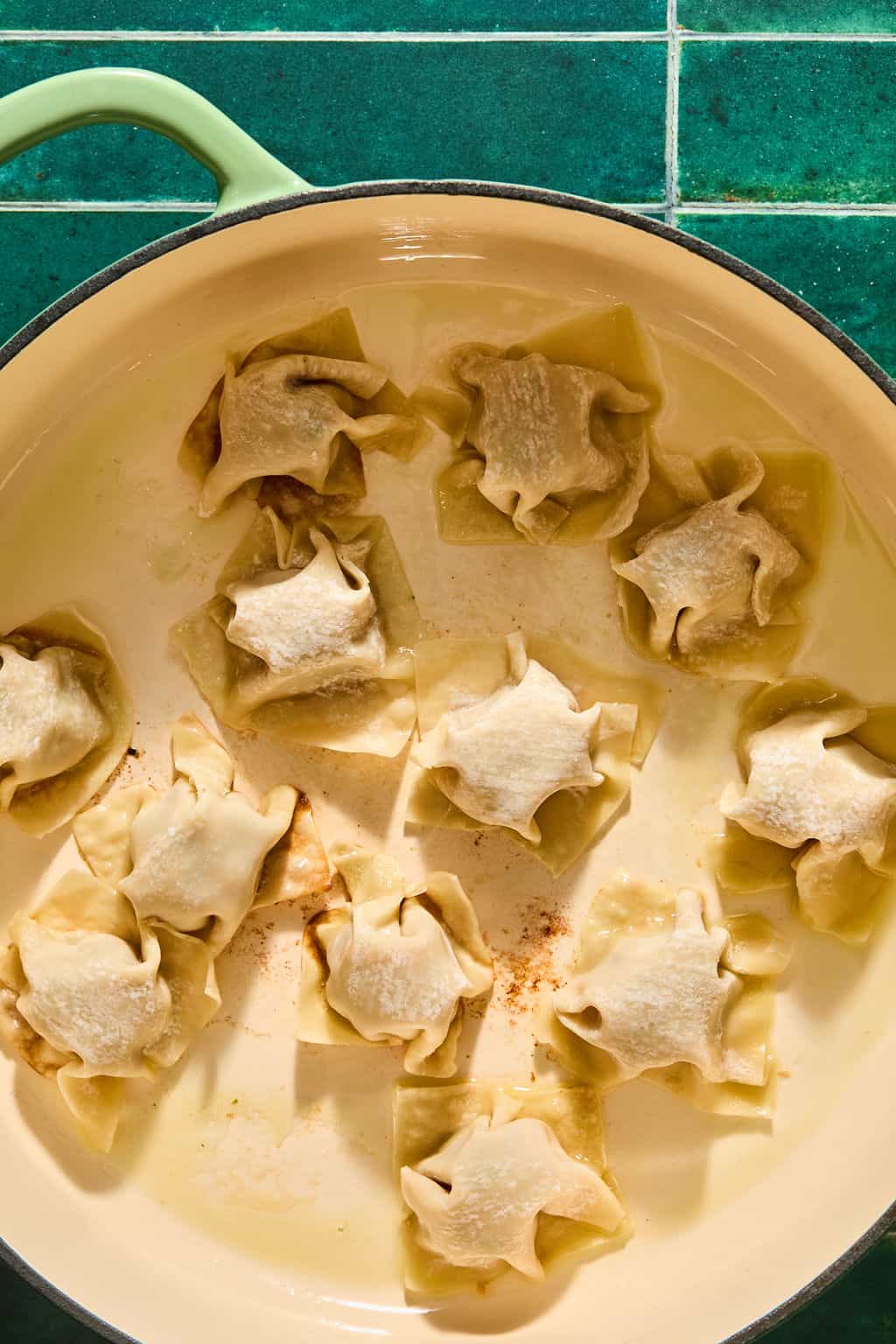 A cream-colored pan with several square wontons arranged inside, each with pleated, pinched tops. The pan sits on a green tiled surface.