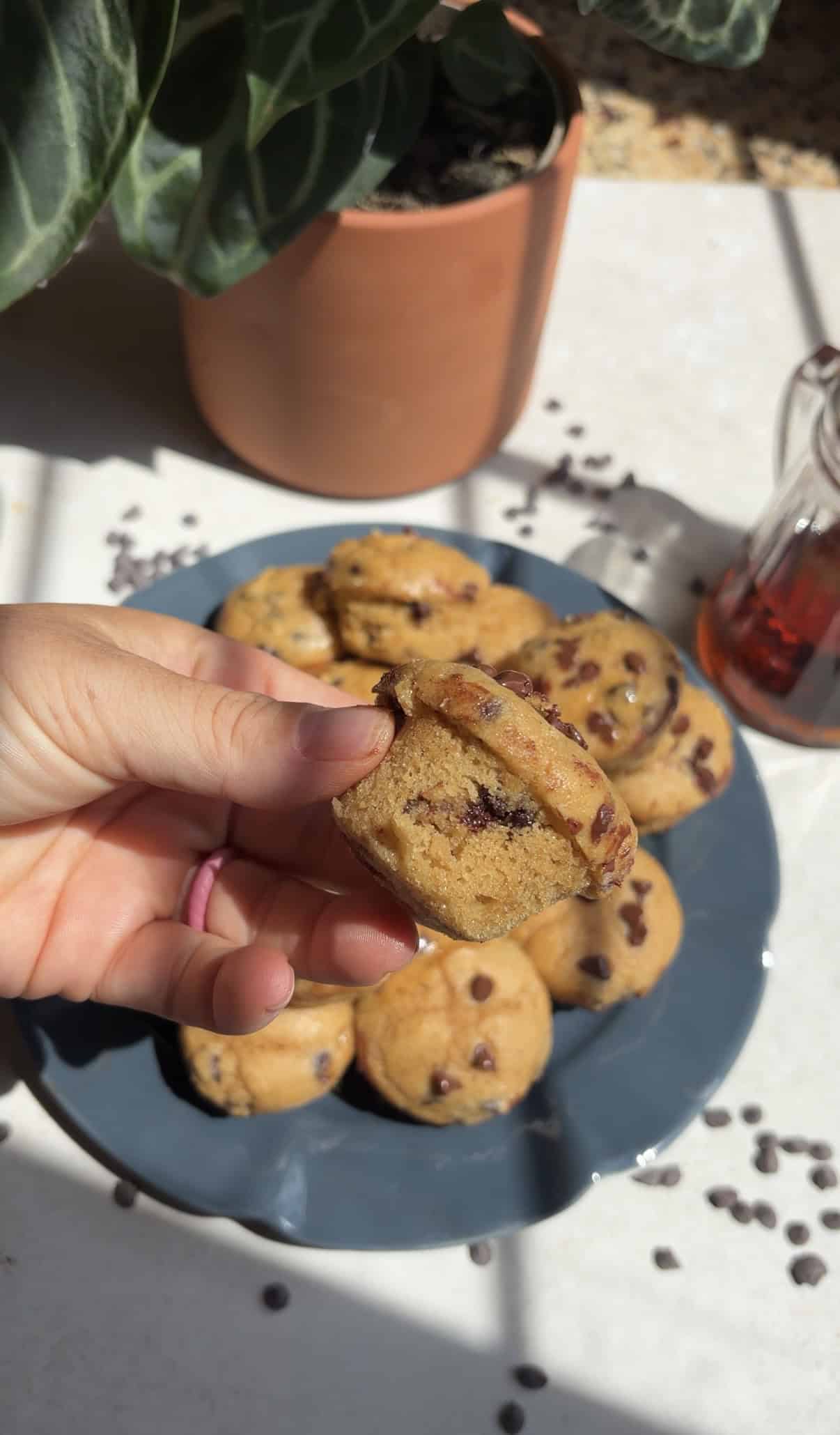 A hand holds a pancake bite above a blue plate with more muffins. Sunlight and chocolate chips are scattered on the white surface.