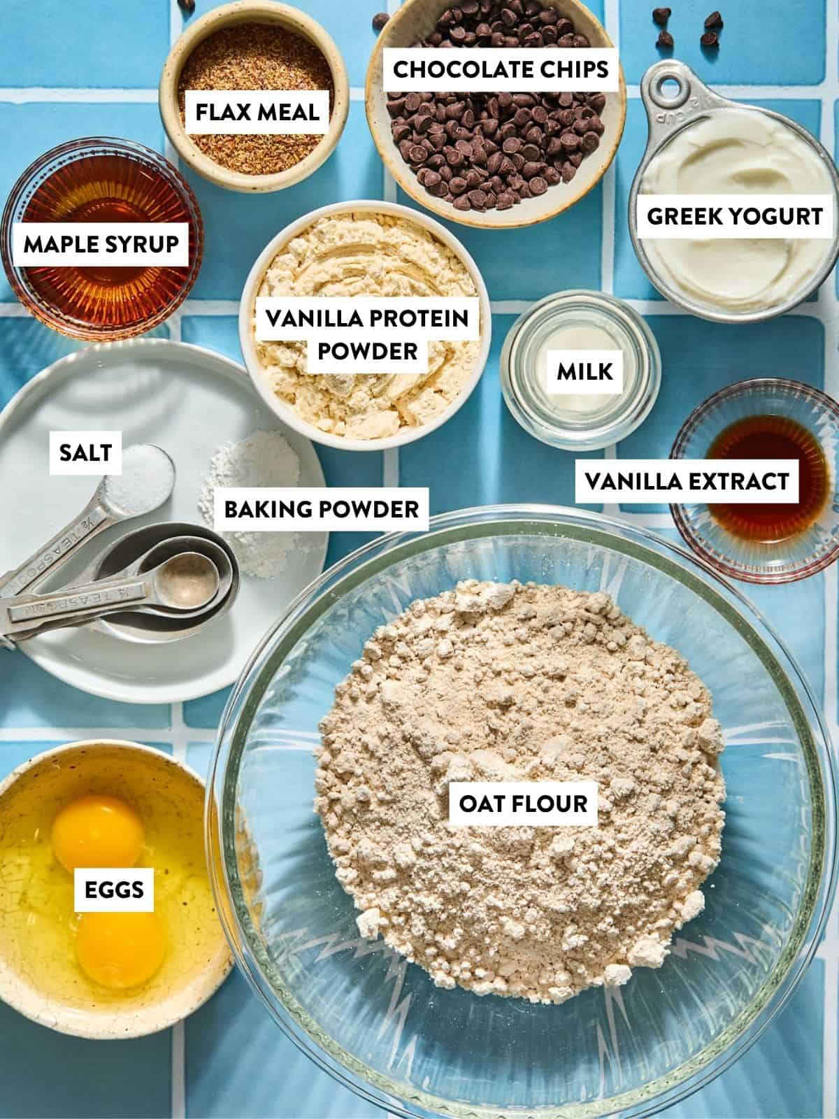 Bowls and measuring cups with labeled ingredients: oat flour, eggs, vanilla protein powder, chocolate chips, flax meal, Greek yogurt, maple syrup, milk, baking powder, vanilla extract, salt, on a blue tiled surface.