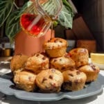 A stack of mini chocolate chip pancake bits on a dark plate, with syrup being poured over them. Theres a green plant and natural sunlight in the background.