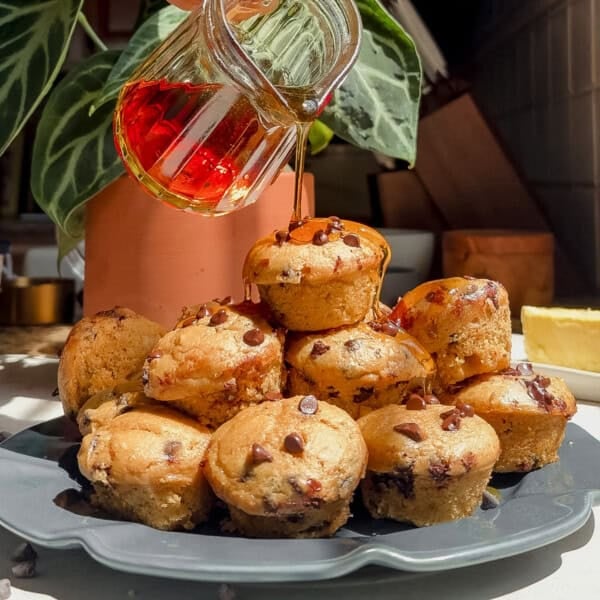 A stack of mini chocolate chip pancake bits on a dark plate, with syrup being poured over them. Theres a green plant and natural sunlight in the background.