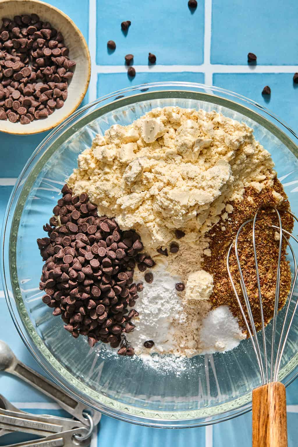 A bowl with chocolate chips, almond flour, flaxseed, baking soda, and salt on a blue tiled surface. Metal measuring spoons and a small bowl of chocolate chips are nearby. A whisk rests in the bowl.