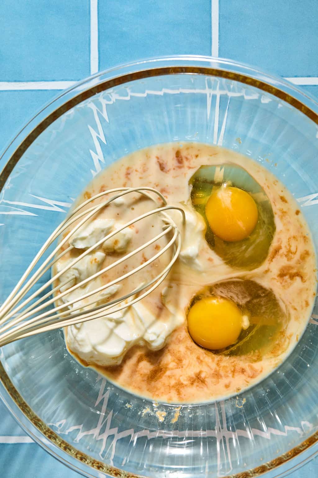 A glass bowl with eggs, greek yogurt, and milk being whisked together on a blue tiled surface.