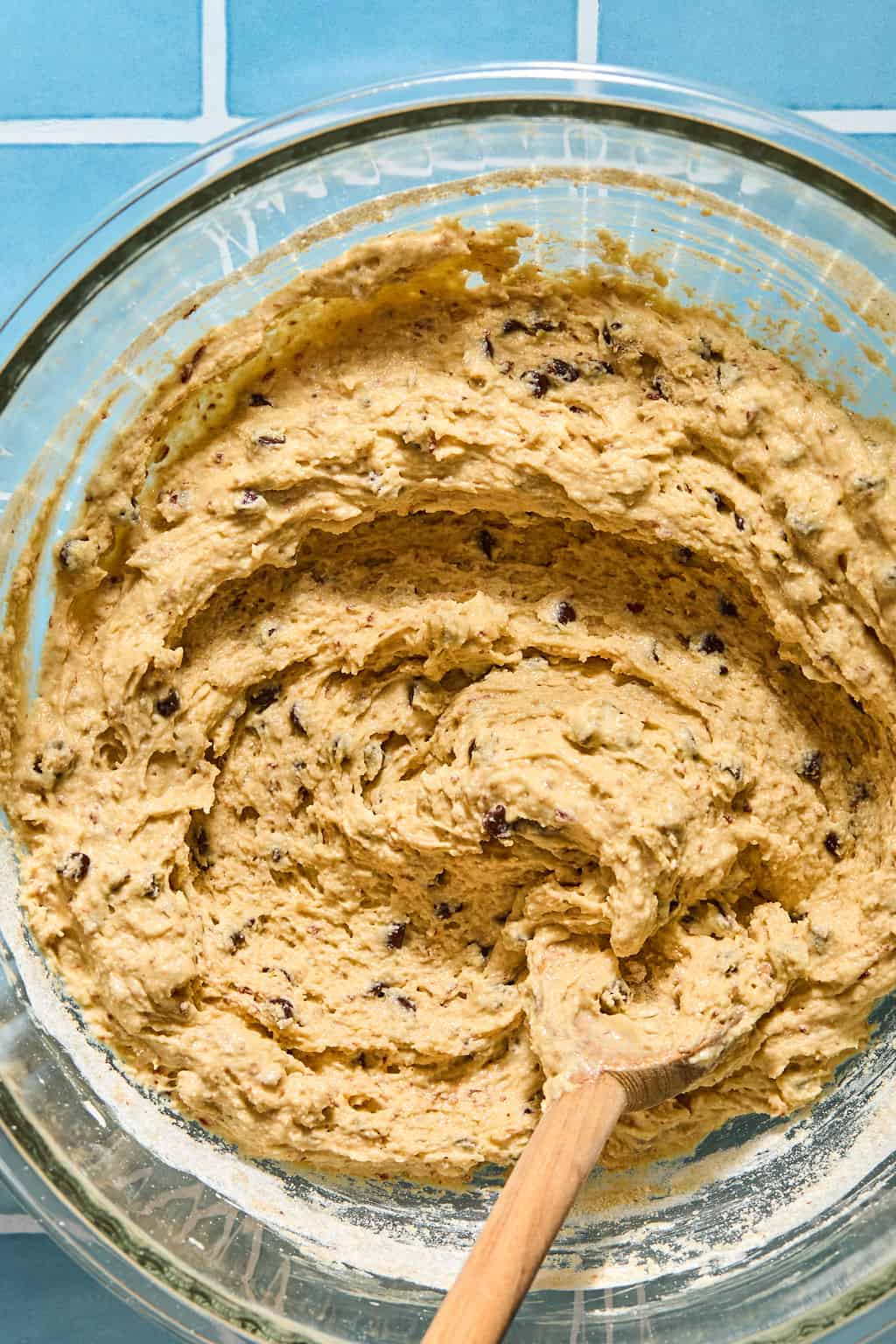 A glass bowl filled with pancake batter is being mixed with a wooden spoon, set on a blue tiled surface.