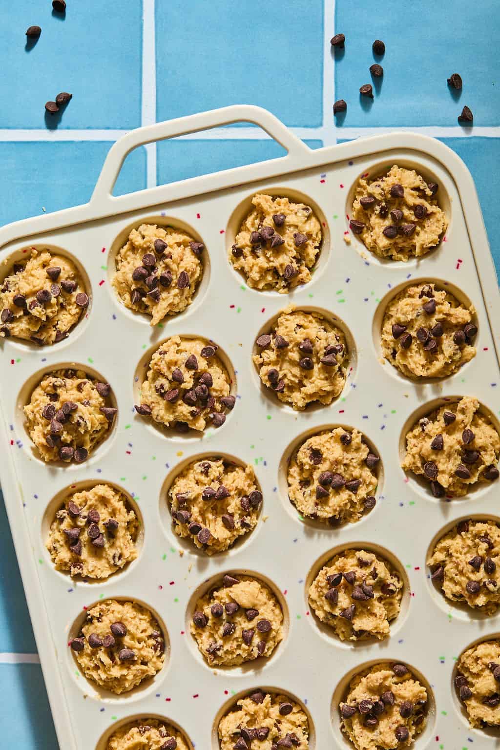 A muffin tin filled with unbaked chocolate chip cookie dough sits on a blue tiled surface, with some chocolate chips scattered nearby.