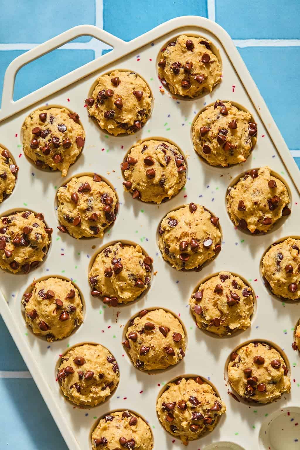 A beige muffin tray holds mini chocolate chip muffins on a blue tiled surface. The muffins are golden brown and topped with chocolate chips.