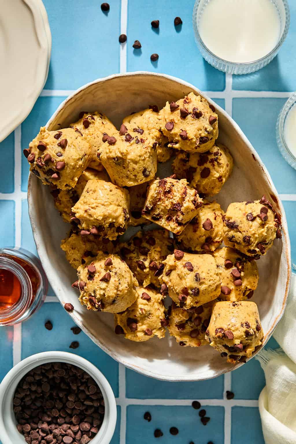 An oval dish filled with chocolate chip mini pancakes on a blue tiled surface, surrounded by bowls of chocolate chips, milk, and syrup.