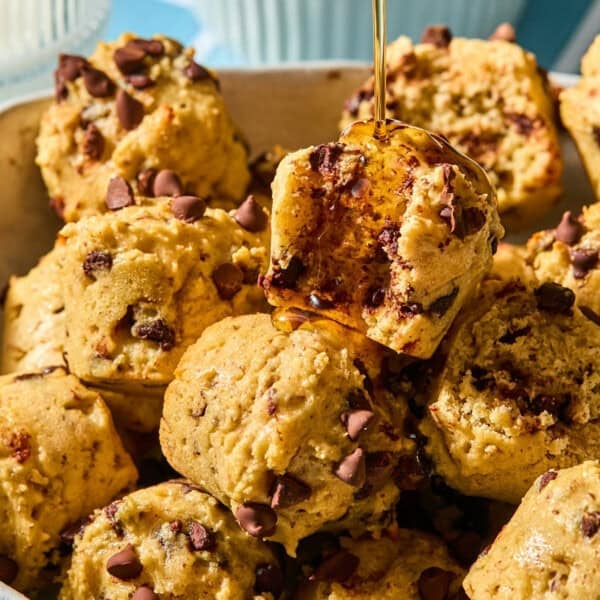 A close-up of chocolate chip muffins stacked together, with honey being drizzled onto a bitten muffin on top. The muffins are golden brown, studded with chocolate chips, and look freshly baked—reminiscent of decadent pancake bites in both size and irresistible appeal.
